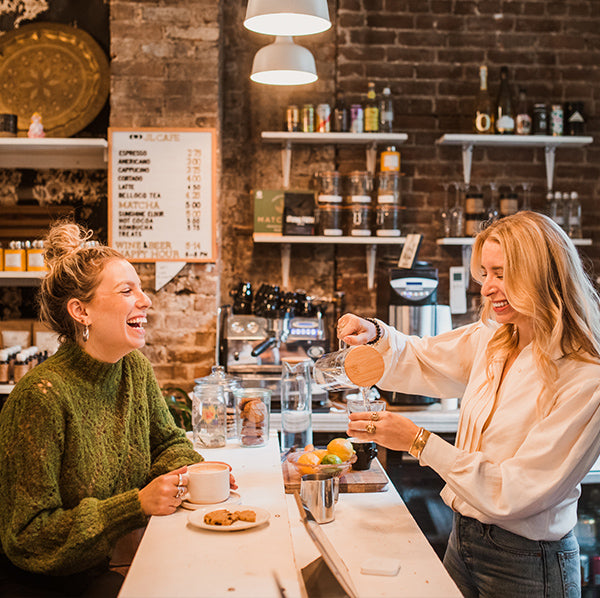 Two women laughing in a small shop with cups of coffee